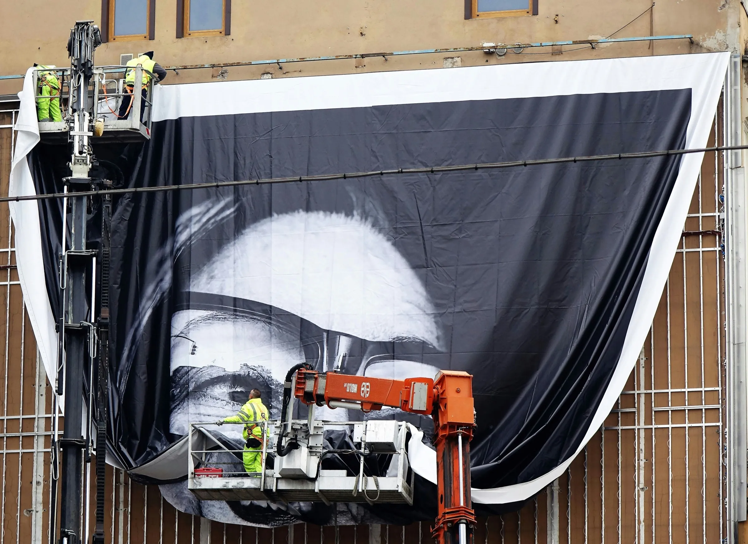 Monteure auf Hubarbeitsbühnen befestigen ein großes bedrucktes Banner an einem Gerüst vor einer Fassade