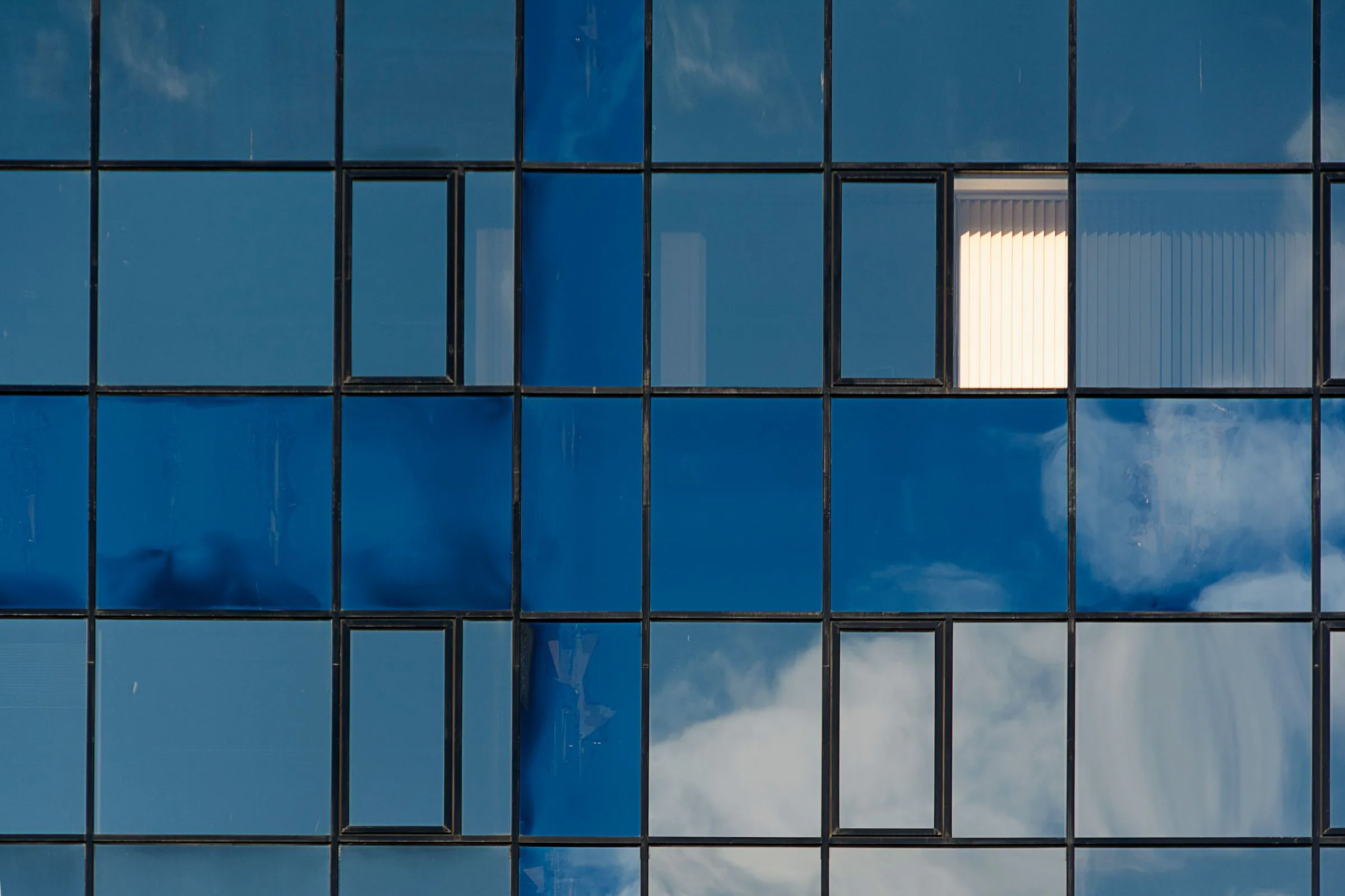 Verspiegelte Fensterfront mit Wolkenspiegelung, ein Fenster lässt die Lamellen dahinter sehen