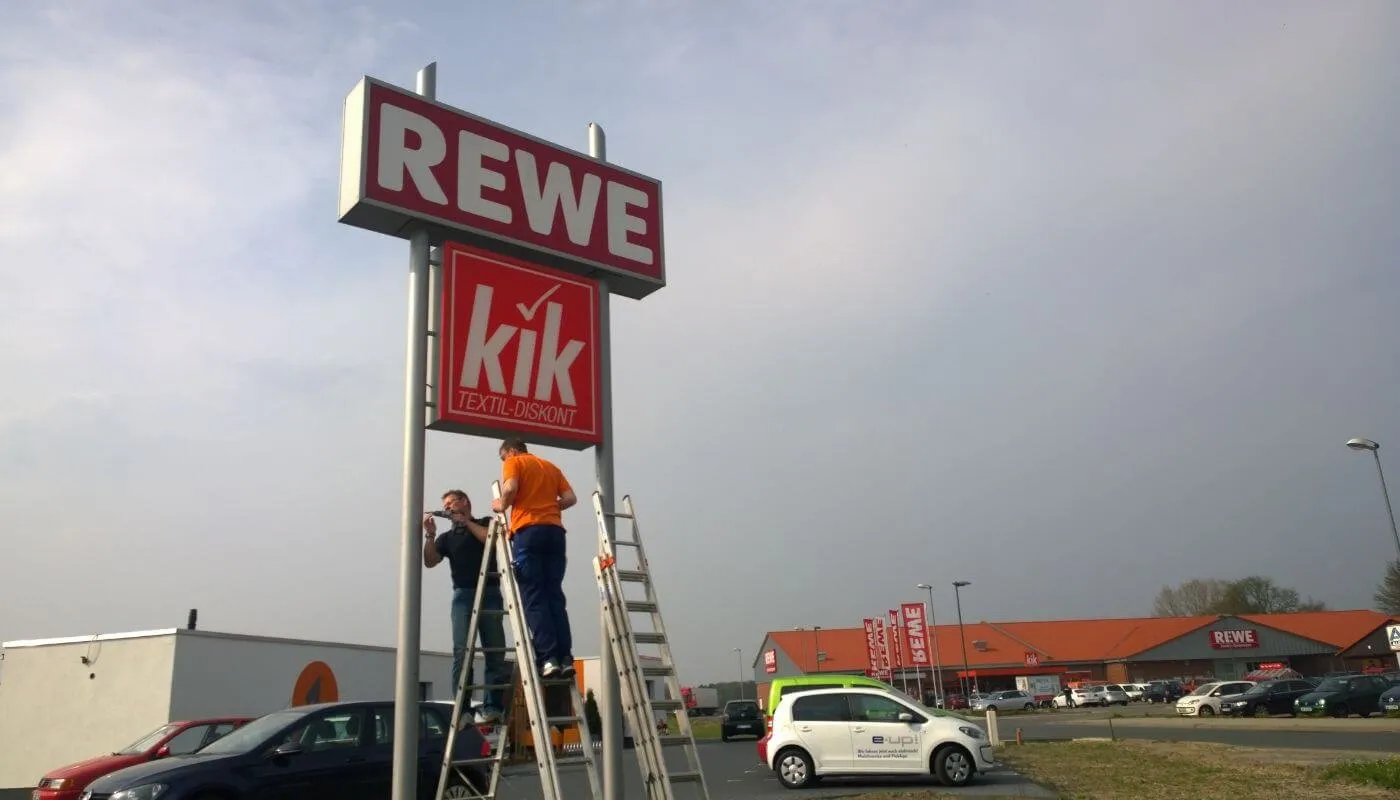 Zwei Monteure von HD Vision auf Leitern bei der Montage an einem Werbepylon vor einem Einkaufsmarkt